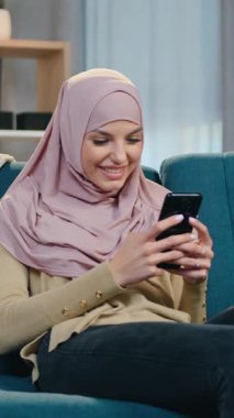 Hoş, pozitif, modern, 30 yaşında, başörtüsü ve modaya uygun giyinen, güzel tasarlanmış bir dairede yumuşak koltukta dinlenen ve telefon uygulamalarına takılan Arap bir kız.