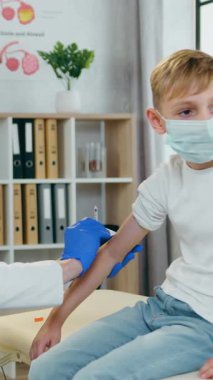 Hoş, yüksek yetenekli, narin, açık renk saçlı, koruyucu maskeli ve steril eldivenli bir kadın doktor. Tıp maskesinin üzerinde oturan genç bir çocuğun koluna iğne yapıyor.