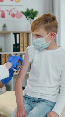 Tıbbi maskeli yakışıklı genç, profesyonel, açık renk saçlı, koruyucu maskeli ve modern tıp dolabında steril mavi eldivenli bir kadın doktor tarafından yapılan iğneden korkuyor.