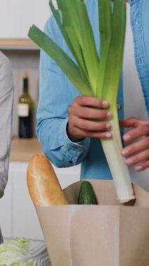 Yakın çekim. Çekici, etnik çeşitliliğe sahip genç bir çift mutfakta birlikte dururken gülümsüyor ve içi taze sebzelerle dolu alışveriş çantalarını açıyor..