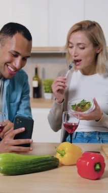 Modern mutfakta telefonu kullanan çok kültürlü genç bir aile. Adam telefonda bir kadına bir şey gösteriyor, kadın gülümsüyor ve salata yiyor. Mutlu aile evde birlikte vakit geçiriyor..