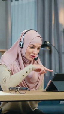 Güzel görünüşlü, gülümseyen, 30 yaşında, türbanlı başarılı bir müslüman kız evde tablet cihazının önünde oturmuş, kulaklıkla video konferansı düzenliyor.