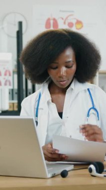 30 yaşında, beyaz önlüklü, video sohbet sırasında bilgisayarın önünde oturan ve önemli notlar yazan 30 'lu yaşlarda başarılı bir Afro-Amerikan kadın doktor.