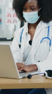 Kendine güveni tam çekici Afro-Amerikan kadın sıhhiyeci koruyucu maske takmış steteskop takmış ve yakındaki tıbbi çalışma odasında çalışan sakallı adam-asistan