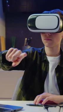 Sanal gerçeklik odaklı gözlüklü beyaz bir çocuk akşam mutfağında oturmuş, VR kulaklığı takıyor. Erkek hareket eden elleri sanal ekranda artırılmış gerçeklik gözlüğü takıyor. Yavaş çekim.