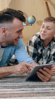 Yakışıklı, neşeli baba ve 10 yaşındaki oğlu evde halıda dinleniyor ve tablet bilgisayardaki farklı videoları, aile eğlencesi konseptini gözden geçiriyorlar.