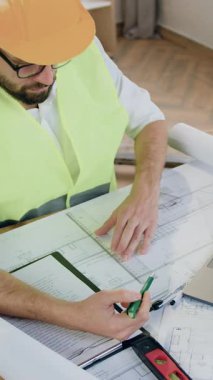 Tasarım ofisinde çalışan iyi görünümlü, kendine güvenen, özel iş kıyafeti ve koruyucu kasklı mühendis ve mimar masada oturuyor ve kağıt planında kalem ve cetvelle çizgiler çiziyor.