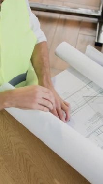 İnşaat endüstrisi konsepti. Güvenilir, kendine güvenen, koruyucu şapka ve yelek giyen uzman sakallı mühendis işyerinde oturuyor ve planlar üzerinde çalışıyor.