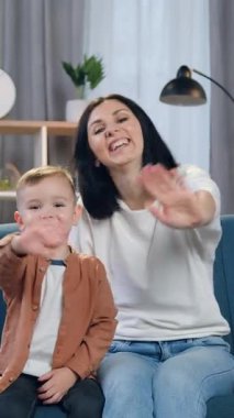 Güzel dekore edilmiş bir odada rahat bir kanepede dinlenen ve herkesi selamlamak için ellerini kameraya sallayan tasasız anne ve 2 yaşındaki neşeli çocuğun portresi.