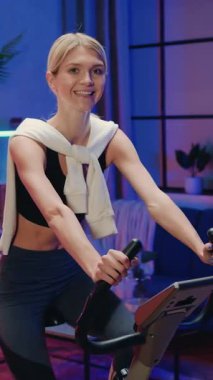 Harika, neşeli, fit atletik, ince sarışın, 25 yaşında, antrenman kıyafetleri içinde. Akşamları bisiklet eğitmeni üzerinde egzersiz yapıyor ve kameraya yansıyor.