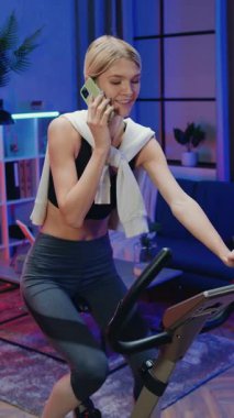 Sağlıklı yaşam tarzı konsepti. Neşeli, sportif, ince, spor kıyafetli, 25 yaşında bir sarışının motosiklet eğitmeni üzerinde egzersiz yaptığı yer. Mobil, keyifli bir gece sohbeti sırasında oda arka planında.