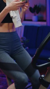 Akşam antrenmanı konsepti. Güzel görünümlü, neşeli, spor kıyafetli zayıf, genç sarışının sabit bisiklet pedallarını çevirdiği ve cep telefonu ve ağır çekim kullanarak mesaja cevap verdiği yer.