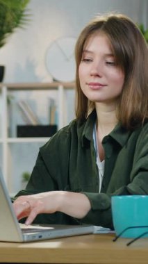 Gülümseyen, kendine güveni tam 25 yaşında, evde çalışmak için özel donanımlı masada oturan ve dizüstü bilgisayarda iyi bir şekilde yazan bir kadın portresi.