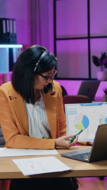 İş dünyası konsepti. Kablosuz kulaklık takan, şık giyinen, internetteki toplantı sırasında dizüstü bilgisayarın önünde oturan ve grafiklerle sunum yapan çekici kararlı iş kadını.