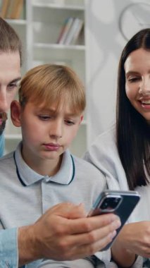 Mutlu, genç bir aile, tatlı baba kız ve oğul anne eli ile kanepede dinlenirken akıllı telefon uygulamasını birlikte oturma odasında, akıllı telefon bağımlılığı ile kullanıyorlar. Yavaş çekim.