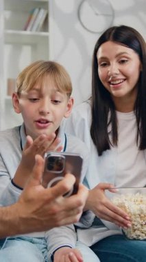 Şirin okul çocukları olan mutlu aile ebeveynleri kanepeye oturup komik akıllı telefonlar kullanarak konferans konuşmaları yapıyor, çevrimiçi içeriği izliyor, gülümsüyor, gülüyor. Yavaş çekim.