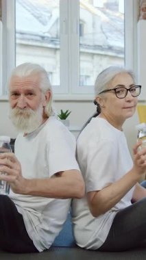 Yakışıklı, pozitif sakallı, 70 yaşında bir adam ve gözlüklü karısı modern oturma odasındaki minderlerde yan yana oturmuş yoga antrenmanında mola verirken kameraya temiz suyla bakıyorlar.
