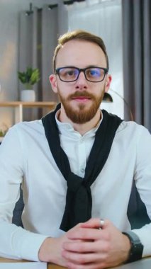 Güzel görünüşlü gülümseyen 25-30 yaşlarında, bakımlı sakallı bir erkek ofis çalışanı portresi. Kameraya jest yaparak bakıyor. Evinde iş yerinde oturuyormuş gibi.