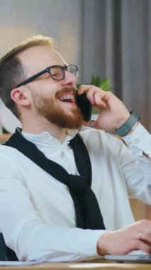 Kendine güveni tam 25-30 yaşında, sakallı bir ofis çalışanı dizüstü bilgisayarın önünde oturuyor ve ev ofisi için özel olarak dekore edilmiş bir yerde güzel bir cep telefonu konuşması yapıyor.