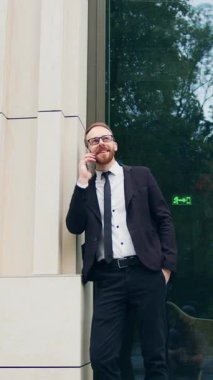 Kendine güvenen ciddi bir adam telefonda konuşuyor profesyonel bir yönetici müşteriyle iletişim kuruyor. İşadamı, modern binanın yakınındaki açık havada akıllı telefondan konuşuyor. Yavaş çekim.