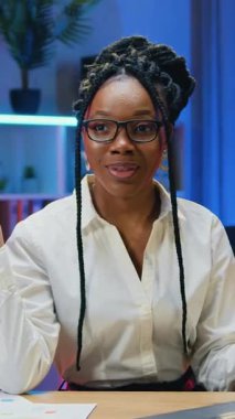 Tatlı, pozitif, başarılı, deneyimli, gözlüklü, beyaz bluz giymiş Amerikan kadın ofis müdürü. Gece ışıklandırılmış çalışma odası arka planındaki çevrimiçi izleyiciler için video kamerada yayınlanıyor.