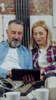 Aile kavramı. Çekici, neşeli, sakallı adam ve güzel sarışın karısının güzel dekore edilmiş bir dairede rahat koltukta dinlenmesi ve tablet cihazındaki uygulamaları düzeltmesi.