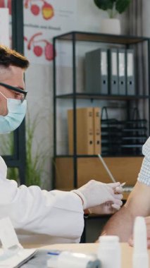 İyi görünümlü, koruyucu maskeli, gri saçlı bir adam aşı yaptırıyor. Salgın hastalık için koronavirüs. Hastane çalışma odasındaki tıbbi maskede uzman bir uzman tarafından veriliyor.