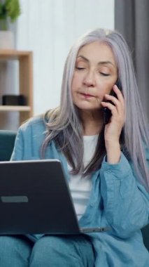 Güzel, konsantre olmuş, olgun, gri saçlı bir bayan güzel dekore edilmiş bir odada rahat bir koltukta oturuyor ve bilgisayar başında yazarken telefonla konuşuyor.