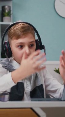 Masadaki kulaklıklı beyaz gömlekli çocuk oturma odasındaki video bağlantısıyla konuşuyor. Evdeyken çevrimiçi ilkokul öğretmenliği dersleri. Yavaş çekim.