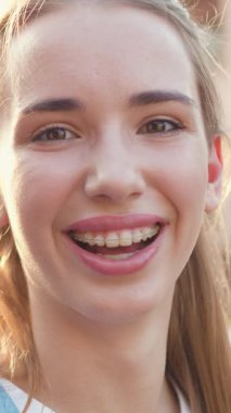 Yakın plan. Genç, güzel bir kız gülümser ve gün batımında hoş bir güneş ışığının arka planında kameraya bakar. Çekici, genç ve şık bir kadının portresi. Yavaş çekim.