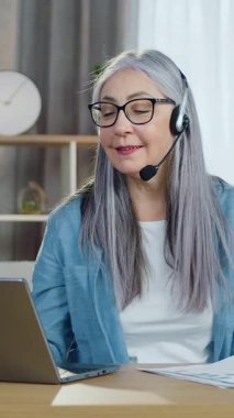 Keyfi yerindeyken sevimli, sakin, 60 yaşında, gri saçlı, kulaklıklı bir kadın video konferansı sırasında dizüstü bilgisayarın önünde oturup müşterilerine rapor sunuyor.