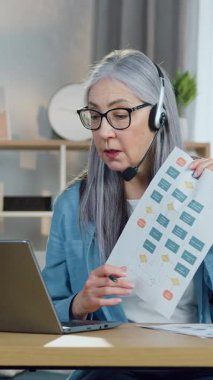 Güzel içerikli, başarılı, kulaklıklı yaşlı bayan çevrimiçi brifing veriyor ve müşterilerle birlikte rapor kullanarak iş yapmanın sonuçlarını analiz ediyor.