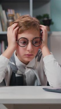 Tired exhausted kid boy leaning his head, with eyeglasses., he is preparing for exams and looking laptop. Boy is doing his homework at the table. Slow motion.