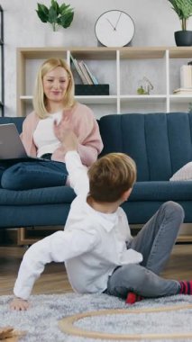 Mükemmel deneyimli, açık renk saçlı bir kadın bilgisayarda yazı yazıyor ve aktif, dost canlısı ergen oğlunu ve kızını araba yarışı yolunda oynarken ve onlara el sallarken izliyor.