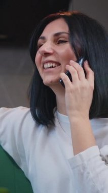 Young woman is sitting at home on the couch and talking on a cell phone. Distance work, working, having conversation with client. Slow motion.