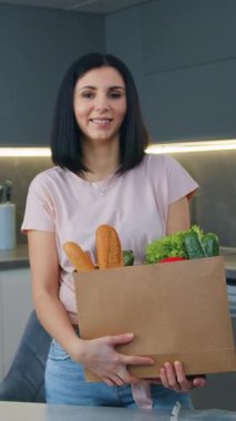 Güzel görünüşlü bir gülümseme, kameraya poz veren 30 yaşında bir esmer. İçinde yeşil bakkaliye ve güzel bir mutfak malzemesi olan kağıt torba ve ekmek var.