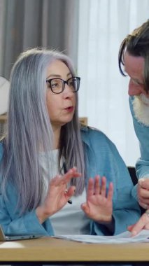 İyi görünümlü, sakin, gri saçlı bir kadın modern oturma odasında dizüstü bilgisayarda çalışıyor ve sakallı kocasıyla keyifli bir şekilde konuşmak için dikkati dağılıyor.