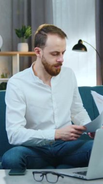 Uzaktan çalışma konsepti. Yakışıklı, başarılı, profesyonel, yetenekli, 30 yaşında, sakallı bir ofis yöneticisi. Çağdaş odadaki yumuşak koltukta oturuyor ve finansal raporlar ve dizüstü bilgisayarla çalışıyor.