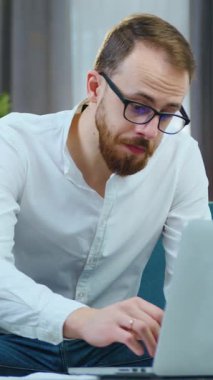 İyi görünümlü, kendine güvenen genç, sakallı bir ofis yöneticisi dizüstü bilgisayarla iş projesi üzerinde çalışıyor ve rahat koltukta otururken farklı belgeler okuyor.