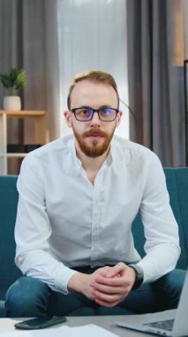 Hoş, sakin, 30 yaşında, güncel giysiler içinde modern bir adam. Çağdaş odadaki yumuşak koltukta oturuyor ve kameraya poz veriyor. Yavaş çekim.