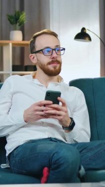 Düşünceli, yakışıklı, pozitif, 25-30 yaşlarında, gözlüklü, güzel tasarlanmış oturma odasında rahat bir koltukta dinlenen ve hareketli, yavaş çekim kullanan bir adam.