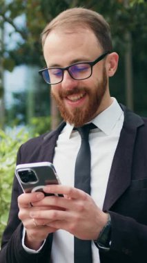 Genç gülümseyen iş adamı, ofis binasının yanındaki bankta otururken akıllı telefon kullanıyor. Erkek girişimci cep telefonunu açık havada eğlenmek için kullanır. Şehirde cep telefonu olan takım elbiseli bir adam. Yavaş çekim.