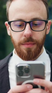 Kapatın. Gözlüklü neşeli adam dışarıda bankta oturmuş akıllı telefon kullanıyor. Ofis binasının yanındaki bankta cep telefonuna mesaj atan kendine güvenen sakallı bir erkek. Yavaş çekim.
