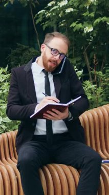 İş adamı telefonda konuşuyor ve defterine bir şeyler yazıyor. Bankta oturan adam cep telefonu kullanıyor. Akıllı telefon üzerinde çalışan yakışıklı bir erkek. Serbest meslek hayatı konsepti. Yavaş çekim.