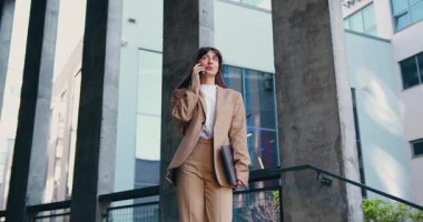 Resmi takım elbiseli kendine güvenen bir iş kadını dizüstü bilgisayar tutuyor ve dışarıda yürürken telefonla konuşuyor. Arka planda modern cam ofis binası. İş iletişimi, önemli çağrı, kurumsal yaşam tarzı, teknoloji, uzaktan çalışma, başarı