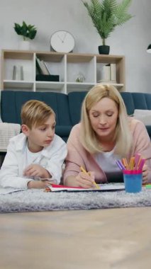 Güzel görünümlü genç erkek ve kız tüylü halıda rahatlarken neşeli, açık renk saçlı anneleriyle ve renkli keçeli kalemlerle kâğıtların üzerine resim çizerken.