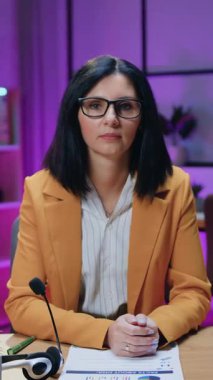 Çekici deneyimli, koyu renk saçlı iş kadını. Gözlüklü ve modaya uygun giyinmiş. Gece vakti kameranın önünde oturuyor.