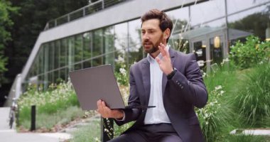 Resmi takım elbiseli odaklanmış bir iş adamının portresi dışarıda kablosuz bir laptopta çevrimiçi konferans düzenliyor. Modern bir cam ofis binasının önünde duruyor, uzaktan çalışıyor ve iş görevlerine konsantre oluyor. Uzak çalışma kavramı