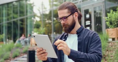 Adamın portresi dışarıda duruyor, dizüstü bilgisayar kullanıyor ve kameraya sıcak bir şekilde gülümsüyor, arka planda modern camdan bir ofis binası var. Kentsel iş ya da yaşam tarzı kavramı.