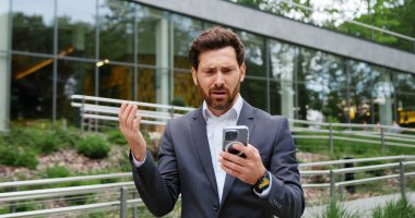 Endişeli iş adamı akıllı bir telefon tutuyor ve bir şehir parkında el kol hareketi yaparken ekrana endişeli endişeli bakıyor. Stres kavramı, acil mesaj, iş iletişimi ve mobil teknoloji.